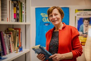 Maureen Corrigan in a red blazer holding a book by a bookshelf