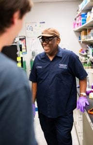 A custodian wearing a navy uniform, a tan baseball cap and purple plastic gloves laughs in a lab with a lab technician.