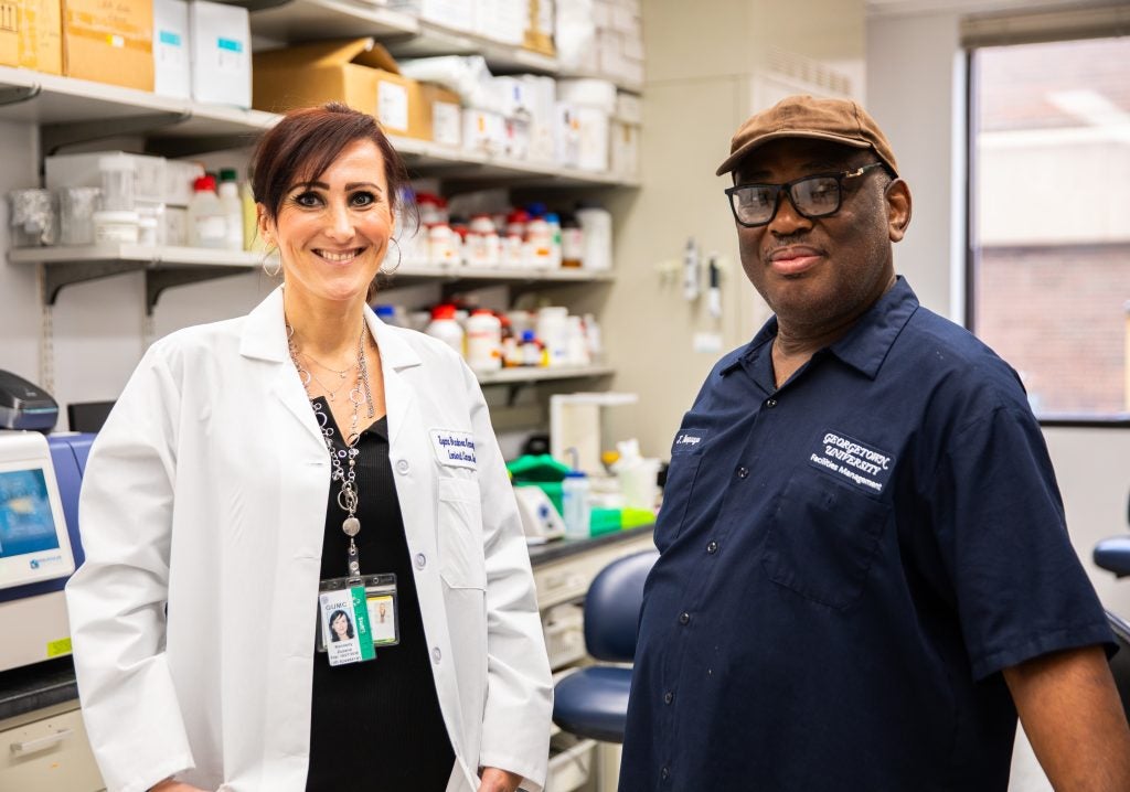 A lab technician wearing a white lab coat stands next to a custodian wearing a navy blue uniform, glasses, and a tan baseball cap.