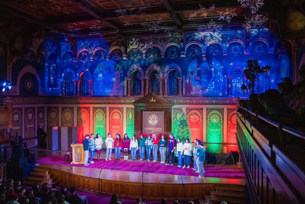 Students sing proudly on stage in Gaston Hall, which is lit in green and red for the holidays