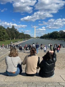 3 girls sit on the steps of the Lincoln Memorial with their backs facing the camera, looking out upon the reflecting pool