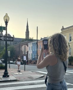 A girl stands at the corner of N and 36th, taking a photo of campus at sunset