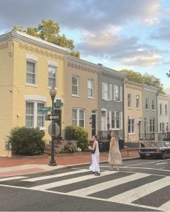 2 girls cross the road in front of Georgetown row homes, smiling at the camera over their shoulder