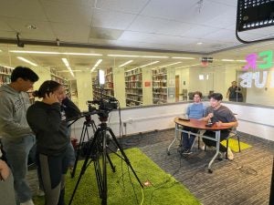 Students practice podcasting, with two students operating cameras and lighting, and another two students being filmed at a desk