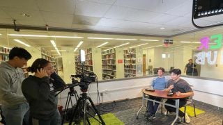 Students practice podcasting, with two students operating cameras and lighting, and another two students being filmed at a desk