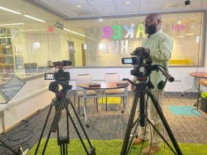 Professor stands in a glass room with podcasting equipment, demonstrating how to work with them