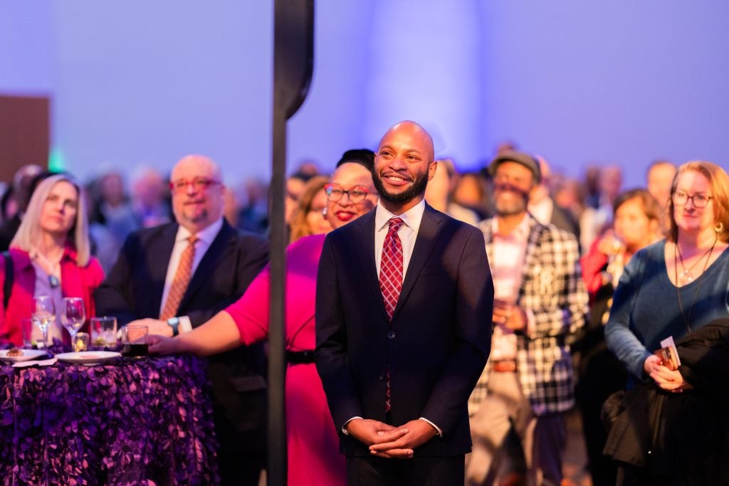 Reginald Douglas, an alumnus of Georgetown, stands in front of a stage and smiles as he is honored with an award. He wears a black suit and a red striped tie, and his arms are clasped in front of him.