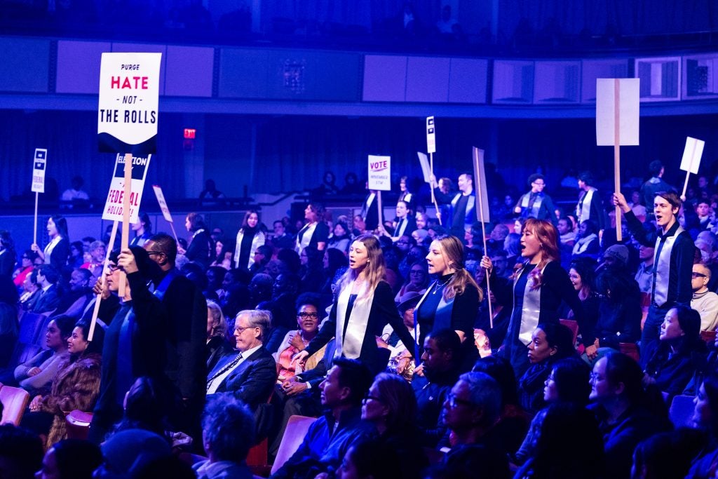 As part of a civic anthem, performers walk toward the stage of the Kennedy Center past audience members carrying signs. 