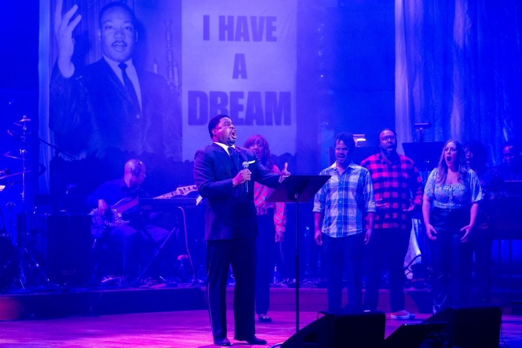A musician sings a song on the stage of the Kennedy Center with other performers and an image of Martin Luther King Jr. and the words, "I Have a Dream," behind him.