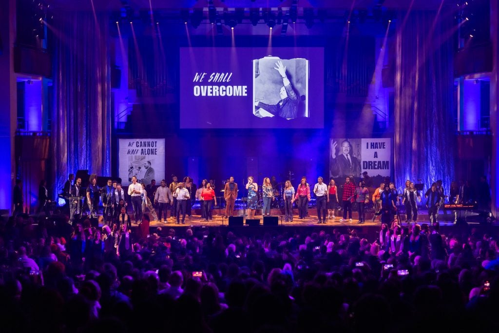 A group of men and women sing on the stage of the Kennedy Center in front of a live audience. Behind them are images of Martin Luther King with the words, "I have a dream"; "We shall overcome"; and "We cannot walk alone."