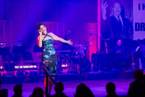 A singer stands on the stage of the Kennedy Center belting out a song and holding out one arm. She's dressed in a blue dress and holds a microphone.