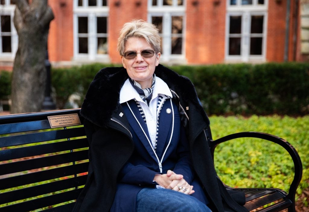A woman with short, cropped blonde hair sits on a bench on Georgetown's campus with her hands clasped on her lap.