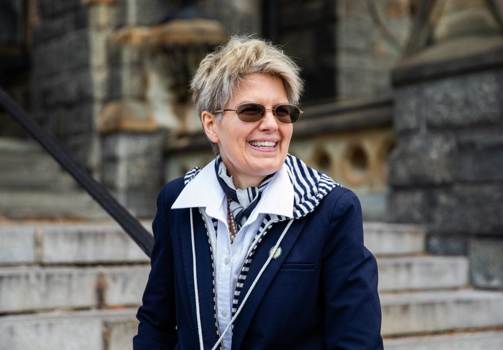 A woman with short, cropped blonde hair wearing sunglasses smiles on the steps of Healy Hall at Georgetown.