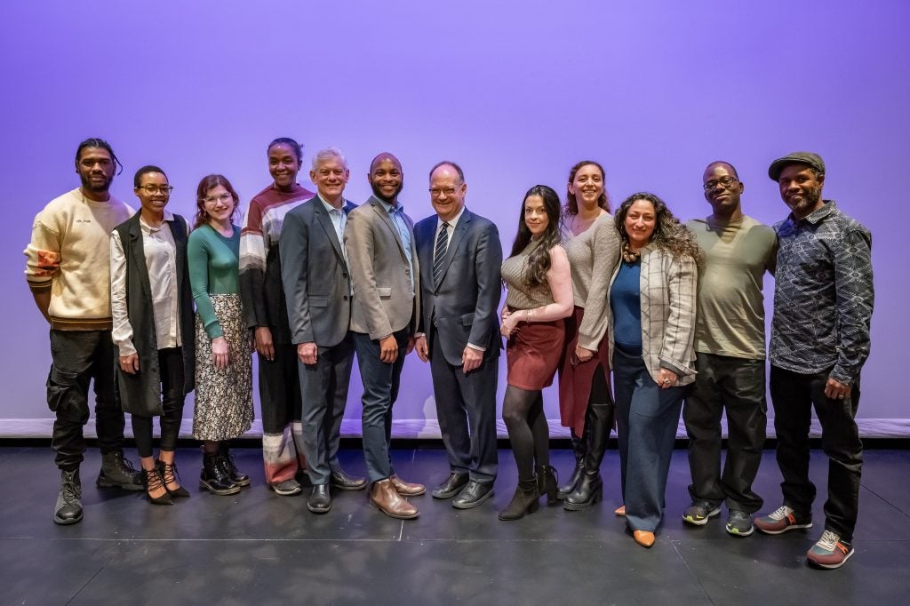 A group of staff members from the Mosaic Theater stand together with Georgetown's president and former basketball coach John Thompson Jr.'s daughter, Tiffany Thompson, in front of a purple background.
