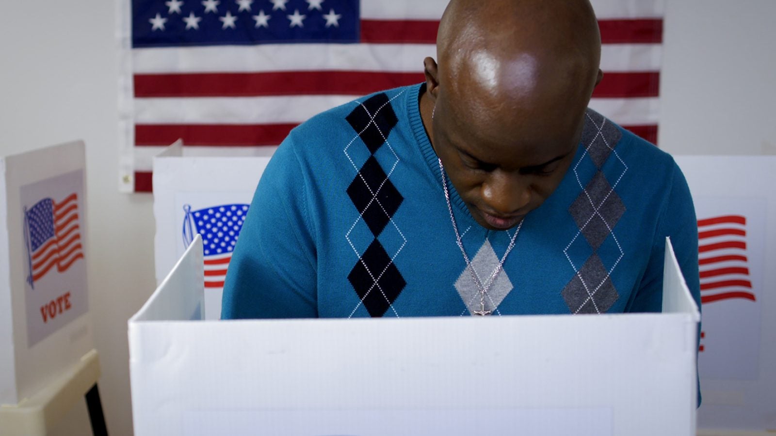 MS front view African American man in blue sweater and wearing a silver cross on a chain casting vote in booth at polling station. US flag on wall in background