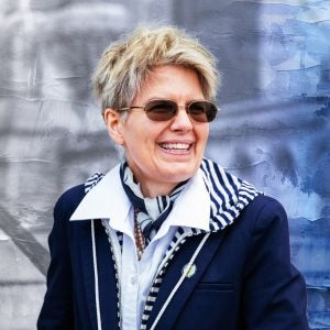 A white woman with short blonde hair smiles and wears a button-up collared shirt with a silk striped scarf and a navy jacket.