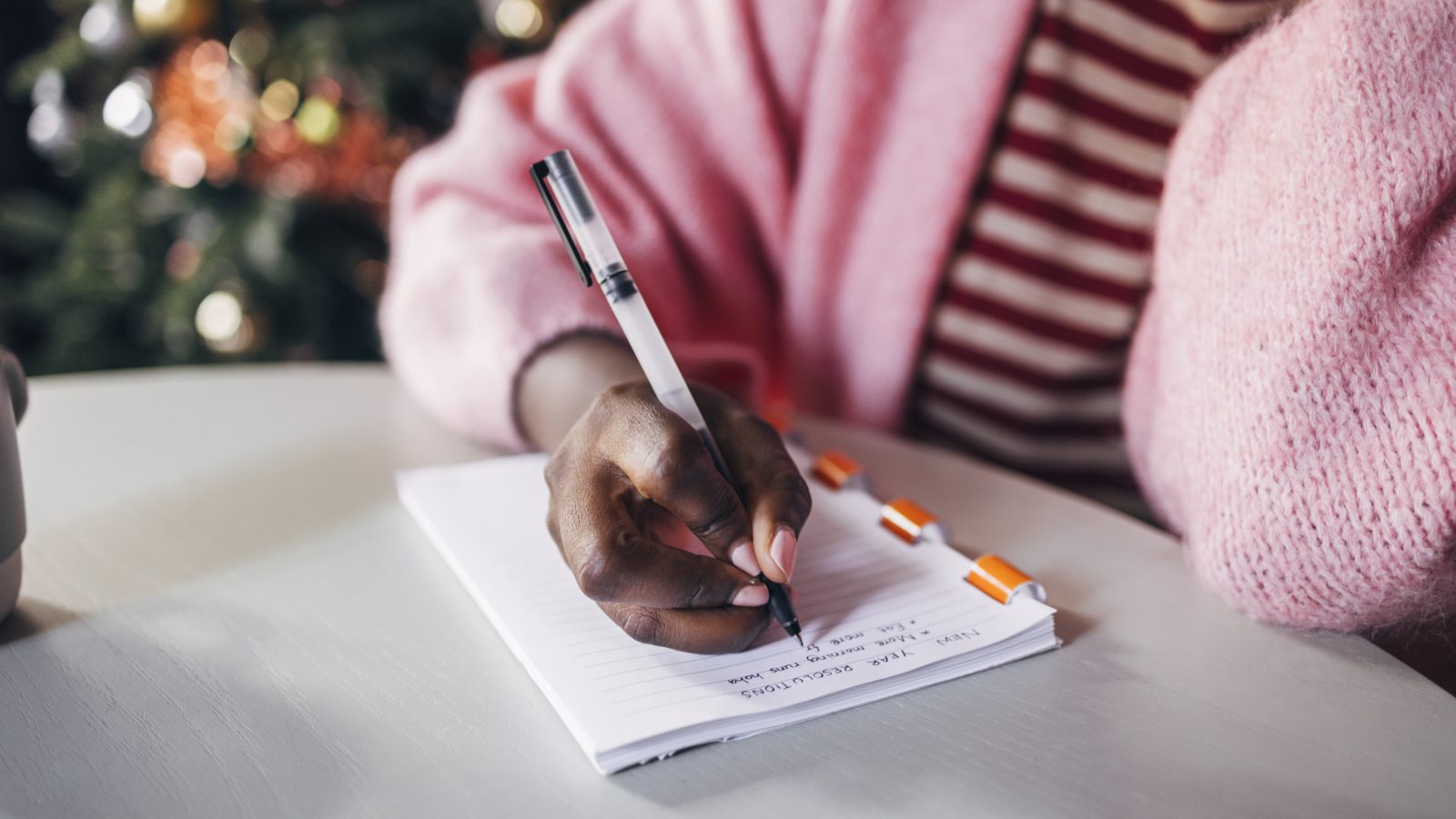 A Black woman wearing a pink sweater and striped pink shirt writes her New Year&#039;s resolutions on a notebook.
