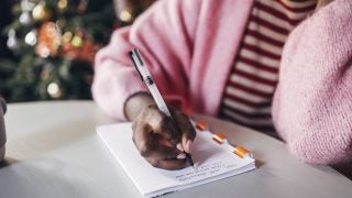 A Black woman wearing a pink sweater and striped pink shirt writes her New Year&#039;s resolutions on a notebook.