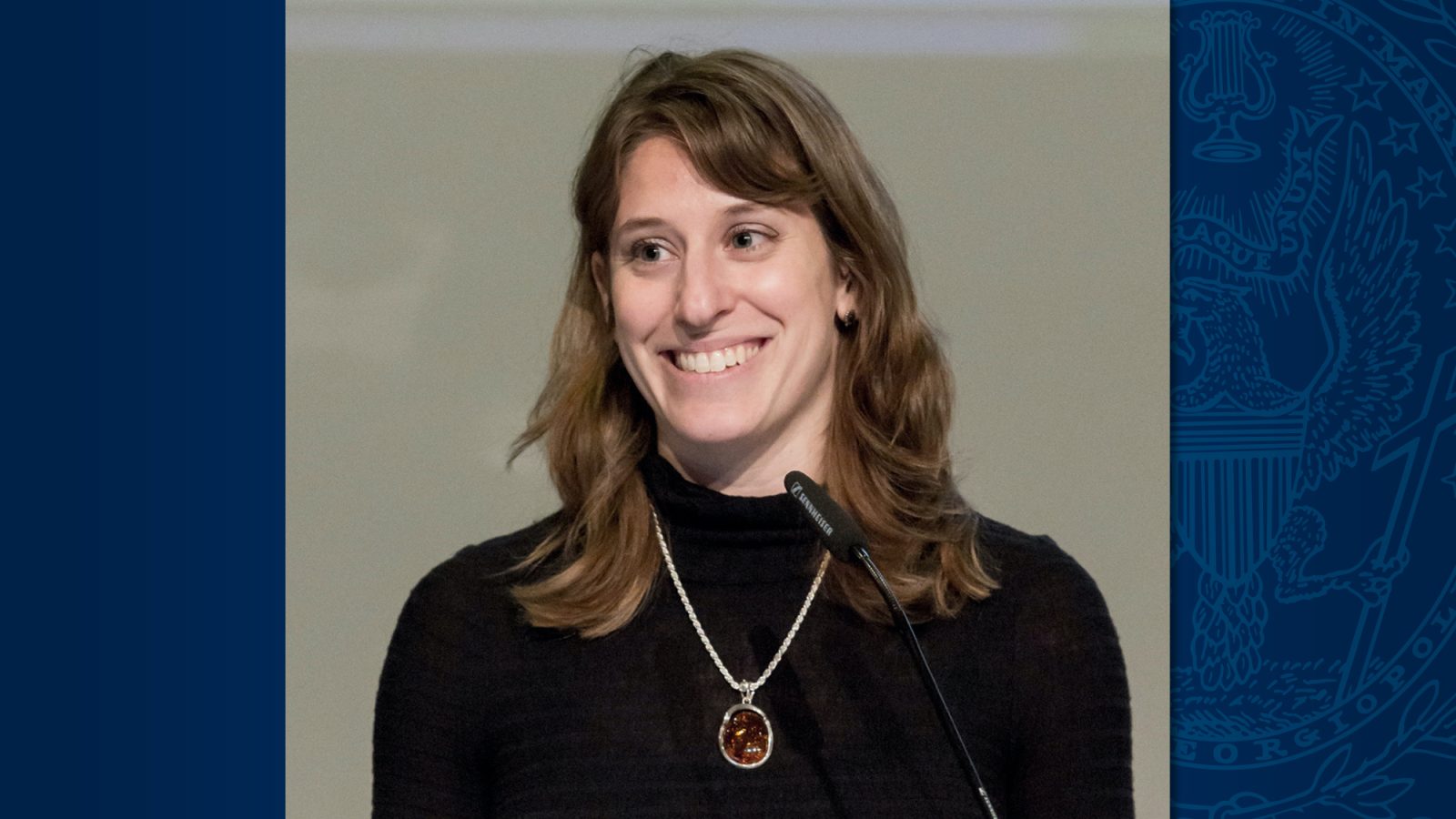 A white woman with brown hair smiles in front of a podium. She wears a black shirt and a necklace.