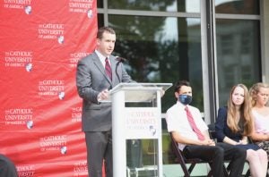 A man speaks at a podium with a banner for Catholic University in the background.