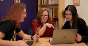 Three woman sit at a table in an office looking at a computer.