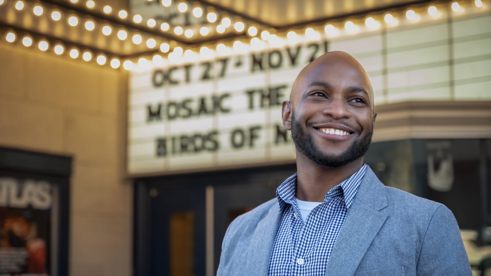 A Black male wearing a button-up shirt smiles with his hands clasped while directing a project.