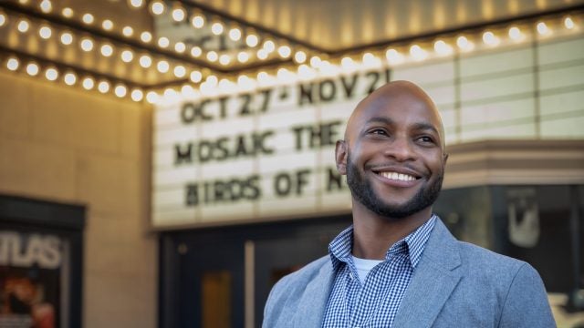 A Black male wearing a button-up shirt smiles with his hands clasped while directing a project.