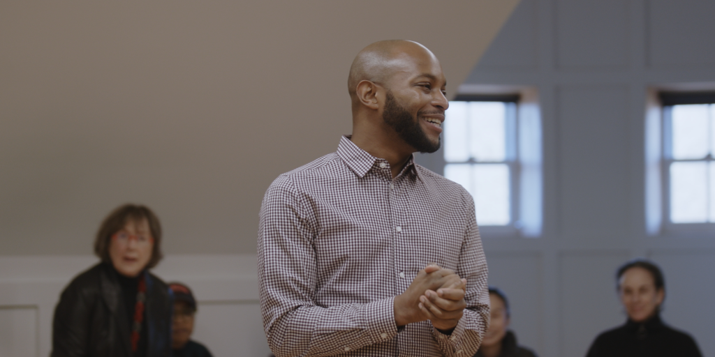 A Black male wearing a button-up shirt and beard smiles and clasps his hands in front of fellow actors and participants as he directs a project.