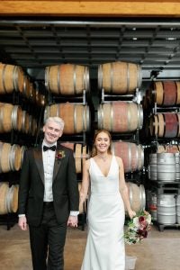 A white couple stands in front of wine barrels on their wedding day. They are holding hands and smiling.