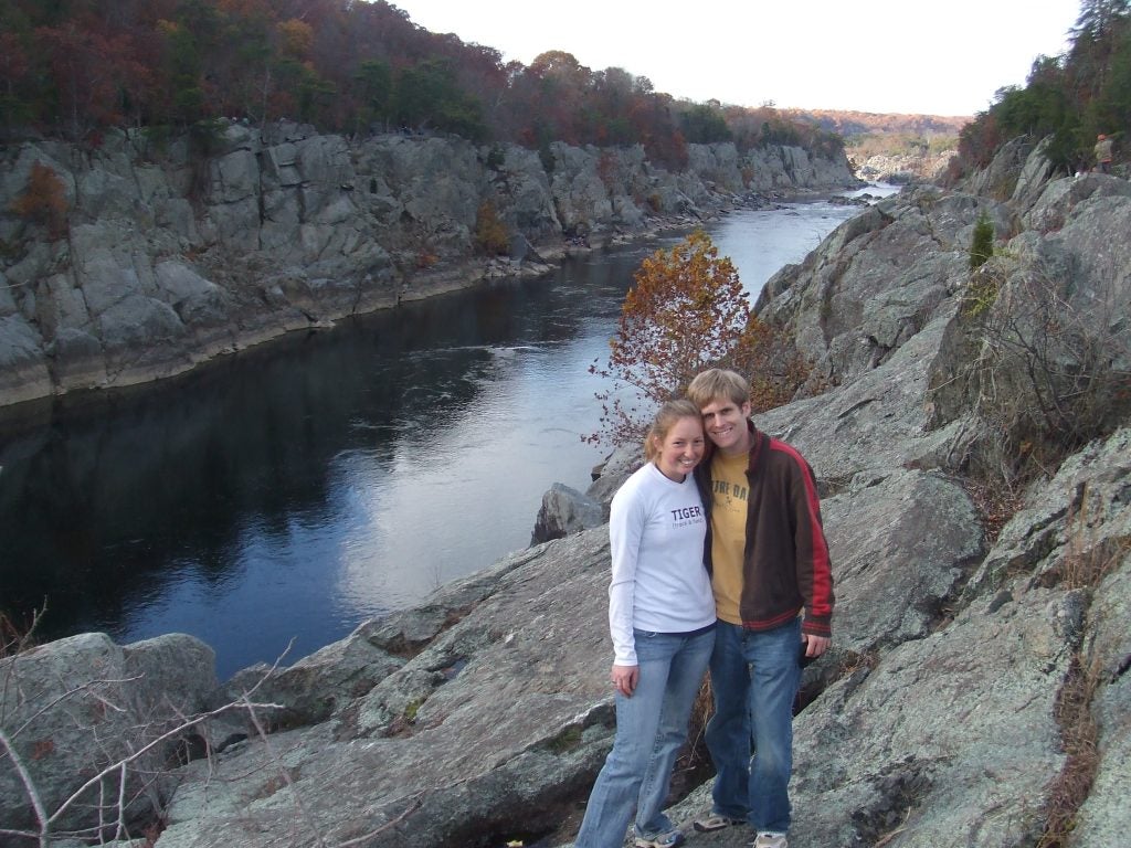 A white man and woman wearing jeans pose on the side of the Potomac River with a rocky background behind them.