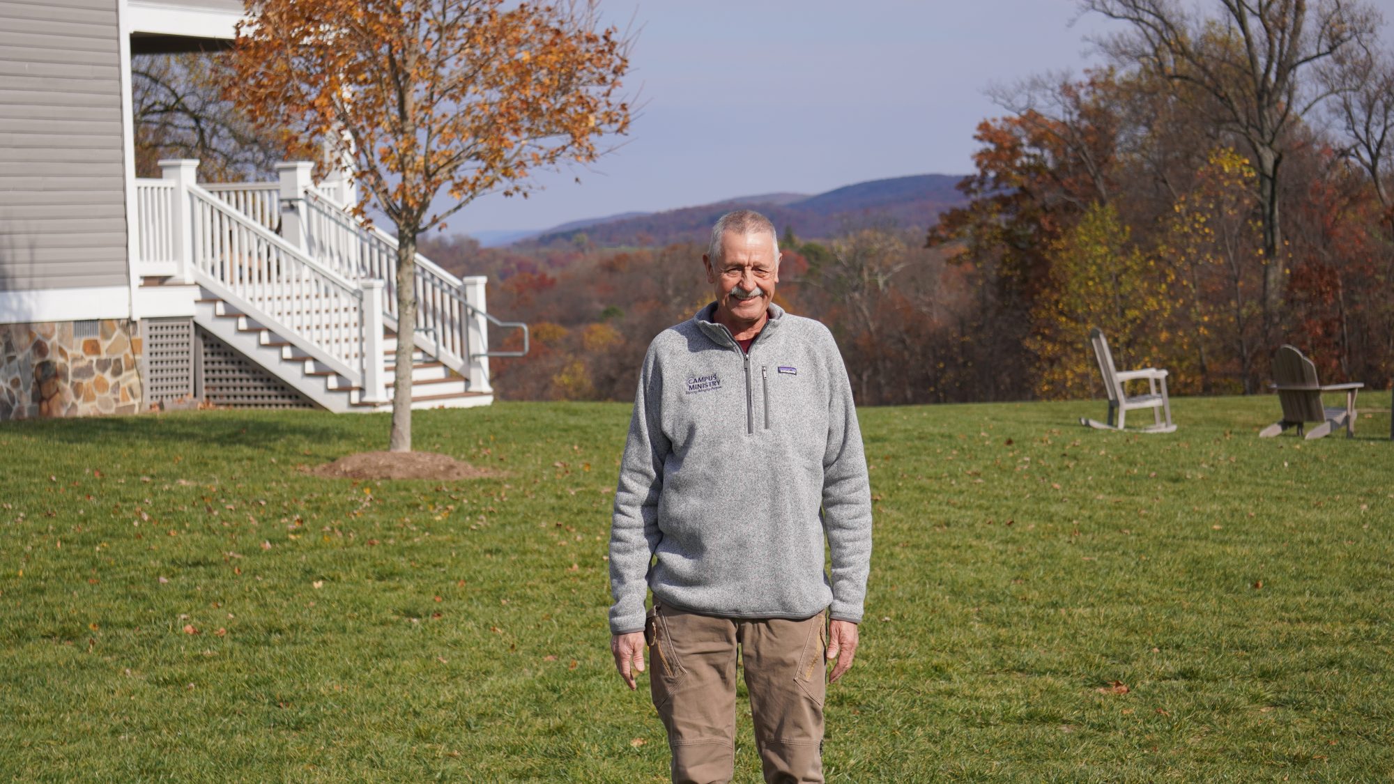 A white man wearing a gray zip-up sweater stands in front of green grass and a mountainous background.