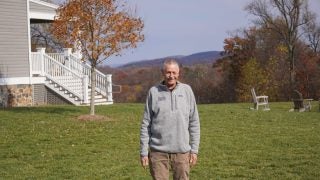 A white man wearing a gray zip-up sweater stands in front of green grass and a mountainous background.