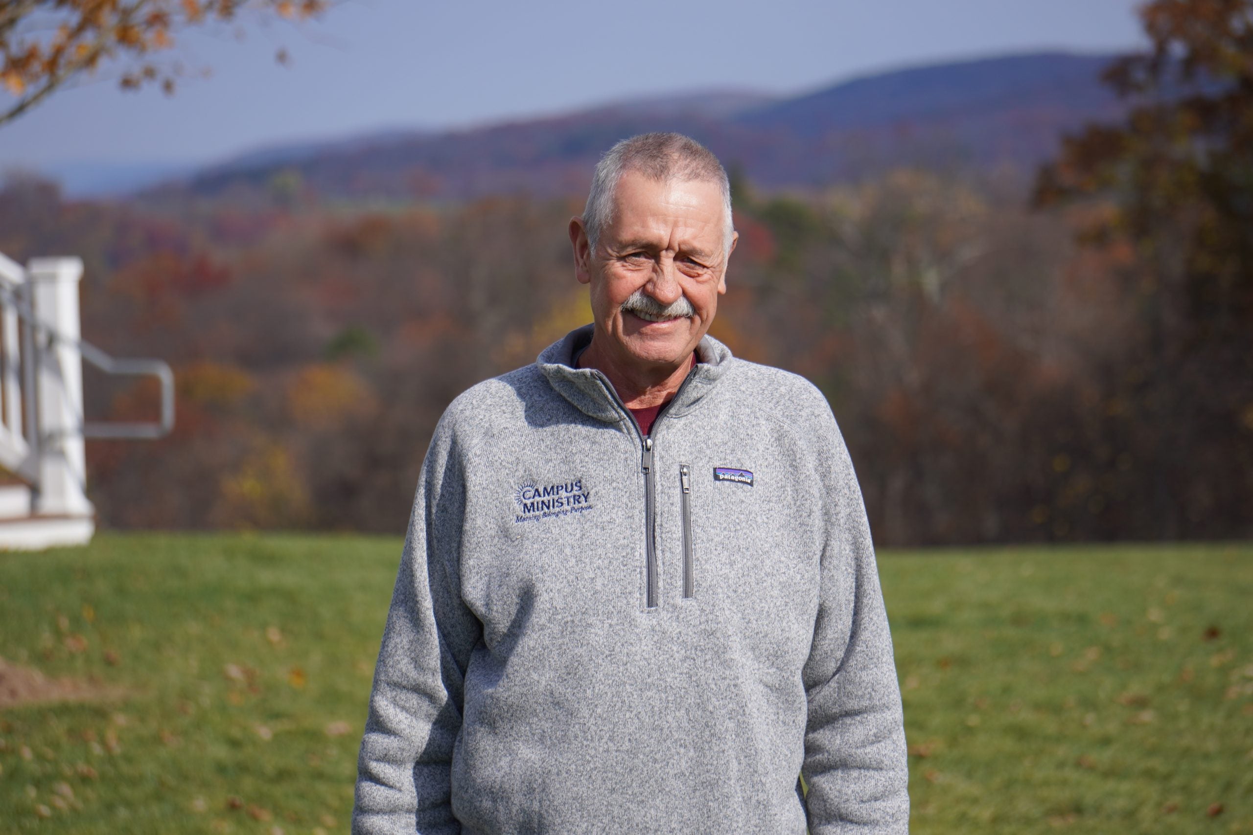 A white man wearing a gray zip-up sweater stands in front of green grass and a mountainous background.