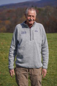 A white man wearing a gray zip-up sweater stands in front of green grass and a mountainous background.