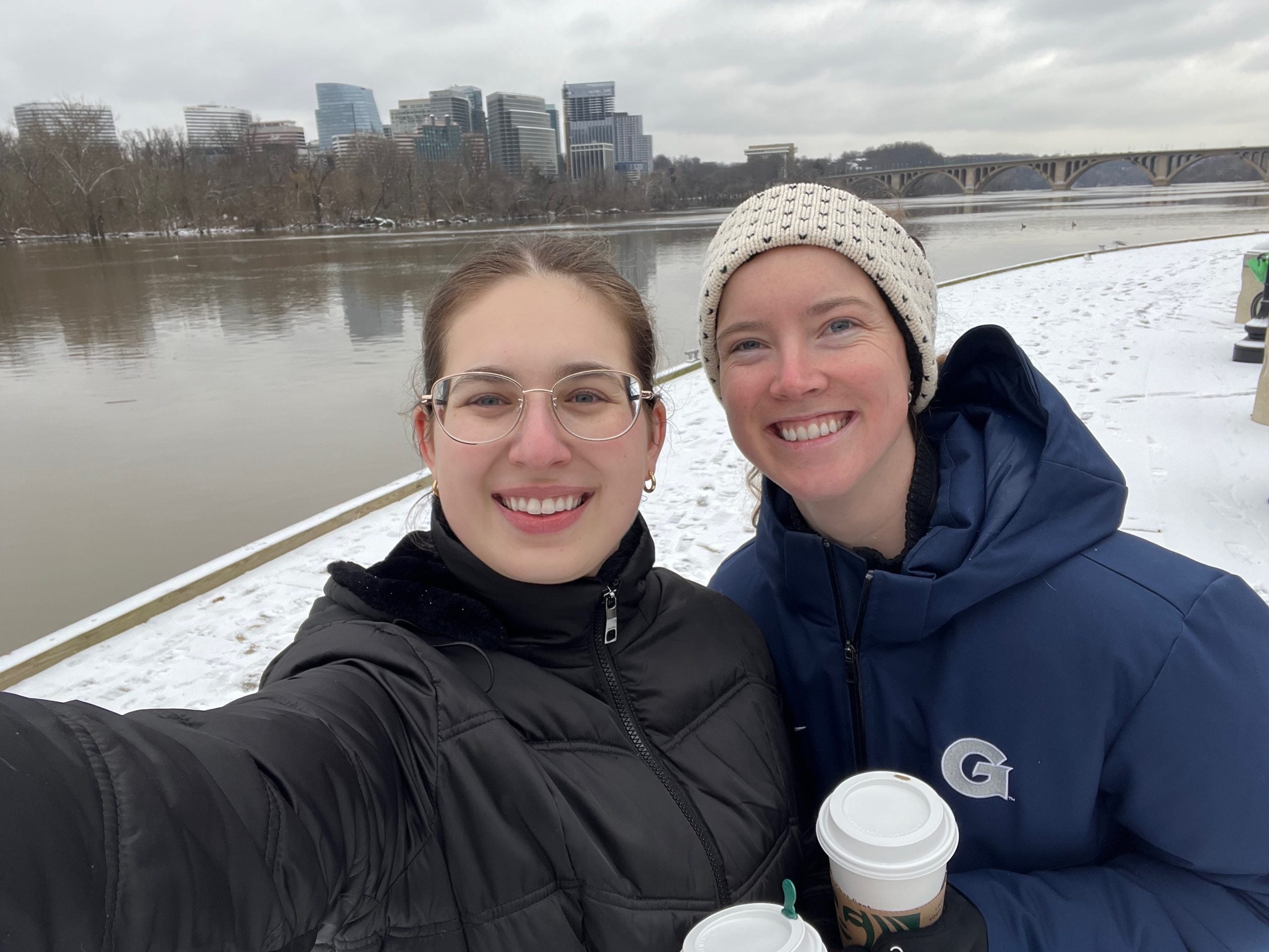 Two white women take a selfie in the winter in front of a river on a cloudy day.