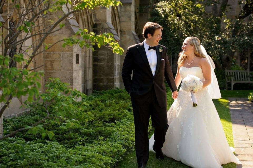 A couple on their wedding day smile at one another as they walk past a church in summer.