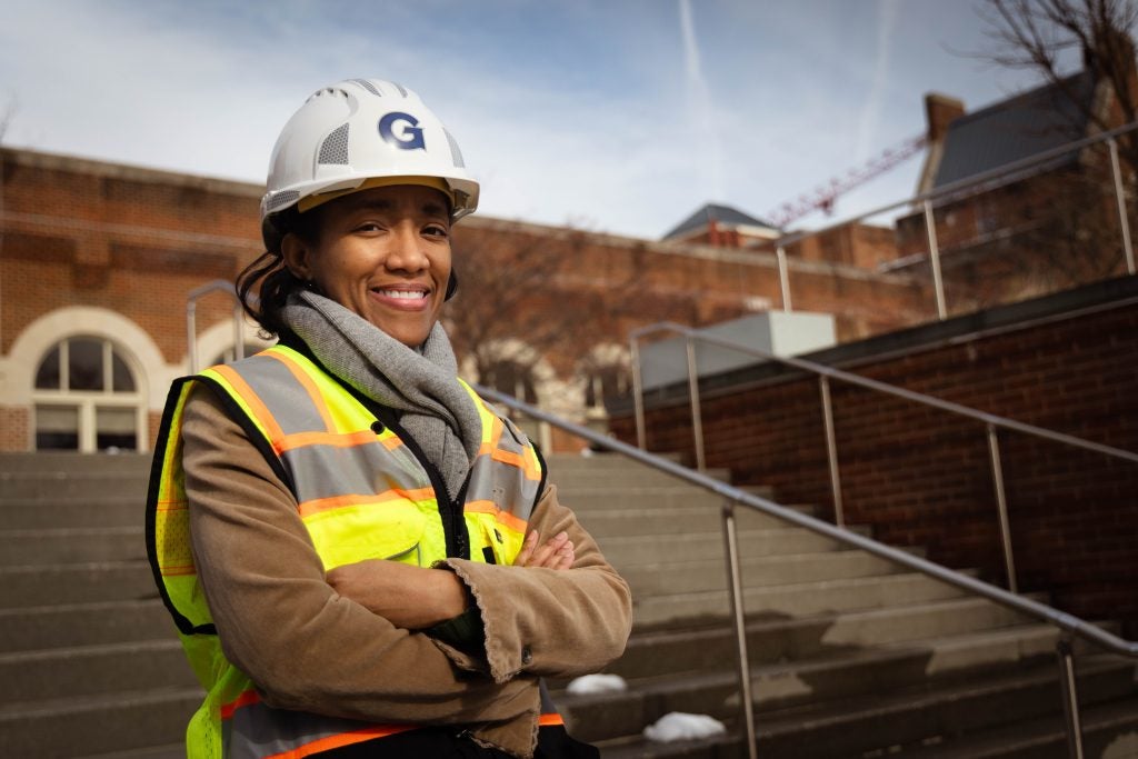 A Black woman wearing a yellow vest smiles while wearing a hard hat with a "G" on the front for Georgetown.