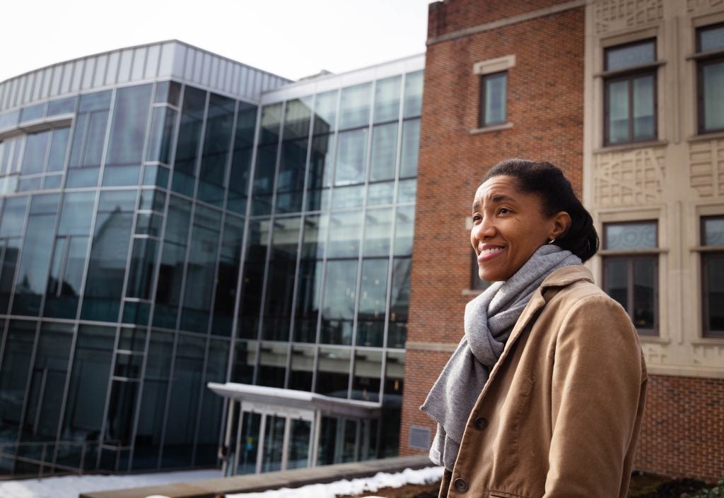 A Black woman wearing a tan jacket and a gray scarf smiles in front of a modern building on Georgetown's campus.
