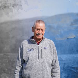 A graphical image of a white man wearing a gray zip-up sweater in front of a blue mountainous background.