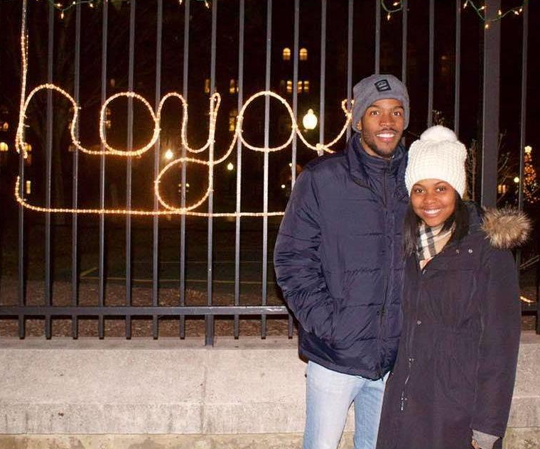 A Black man and woman pose together in the winter outside the Georgetown gates where a "Hoya" Christmas decoration lights up behind them.