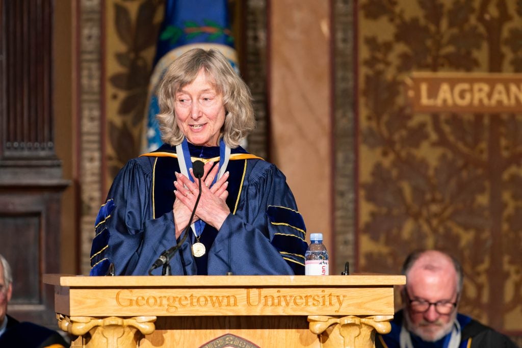 Deborah Tannen, a Distinguished University Professor, wears academic robes while standing with her hands clasped to her heart behind a podium.