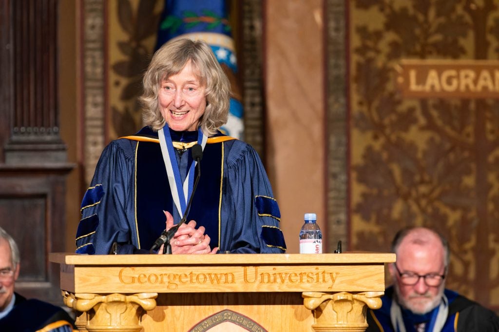 Deborah Tannen, a Distinguished University Professor, wears academic robes while standing with her hands clasped behind a podium.