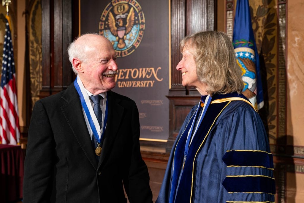 A husband and wife smile at each other on stage. The husband wears a suit and a medal and the wife wears an academic robe and a medal.