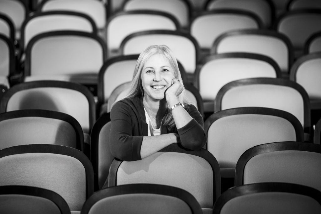 A black-and-white image of a white woman sitting in an empty event space. She leans over the seat in front of her and smiles.