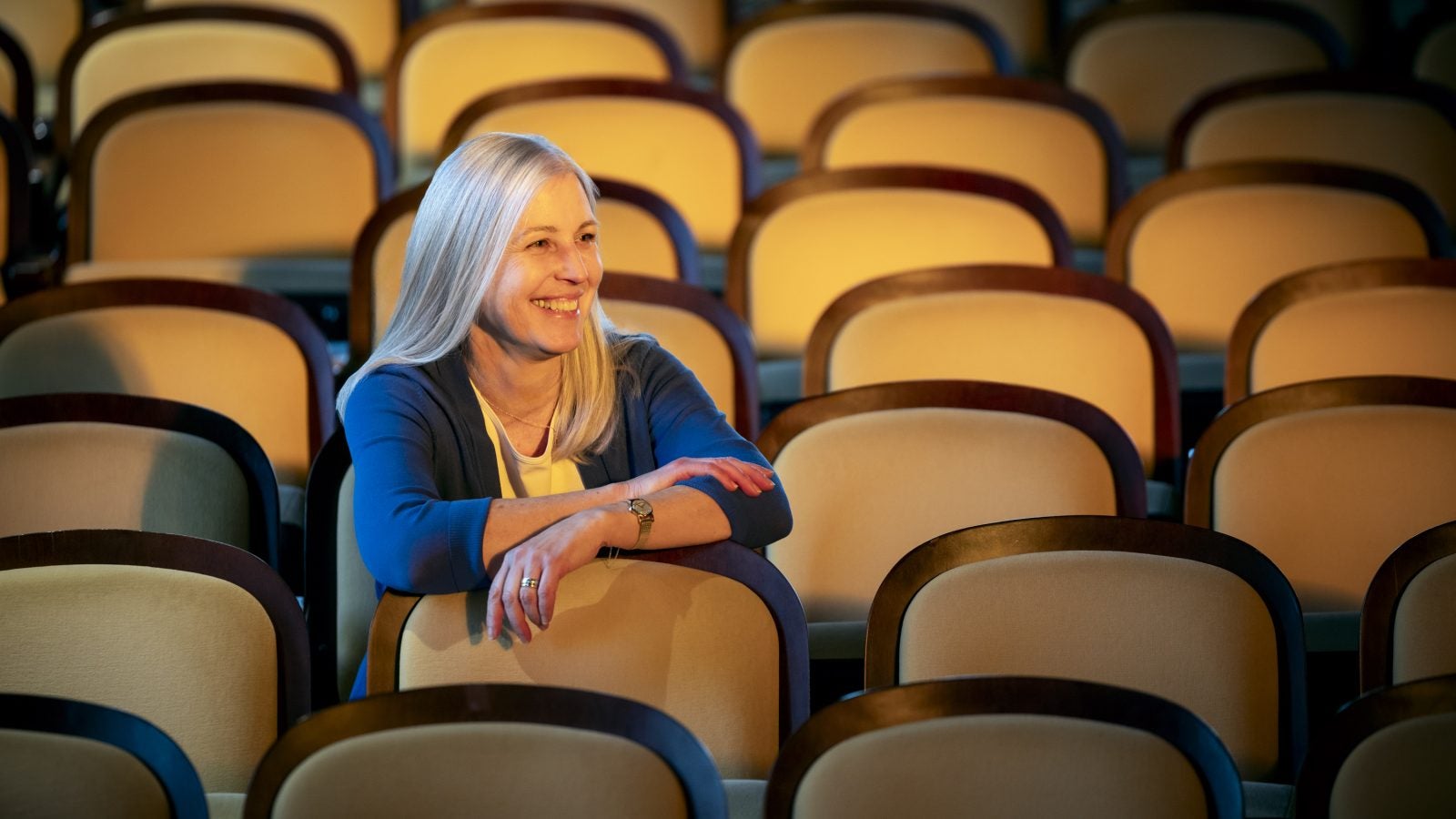 A white woman sits in an empty event space. She smiles with her arms over the seat in front of her.