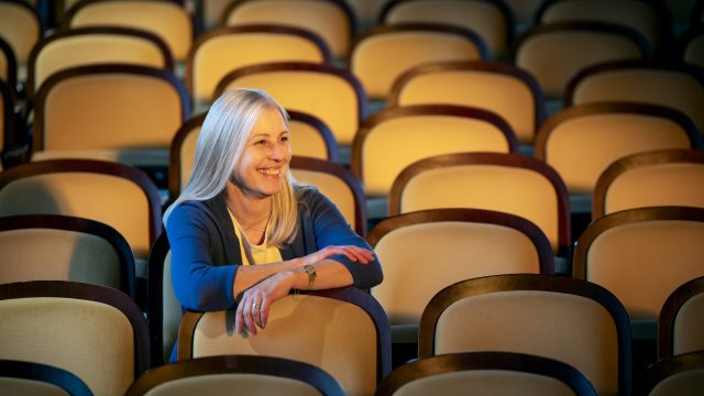 A white woman sits in an empty event space. She smiles with her arms over the seat in front of her.