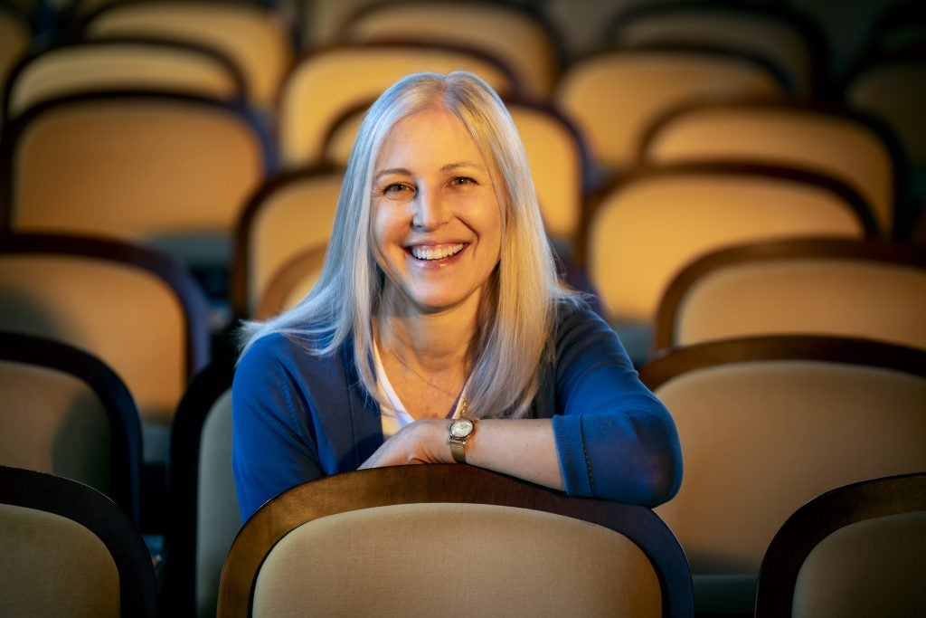 A white woman sits in an empty event space. She smiles with her arms over the seat in front of her.