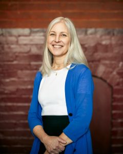 A white woman with gray hair smiles in front of a red brick background at Georgetown. She wears a white shirt, a blue cardigan and a black skirt.