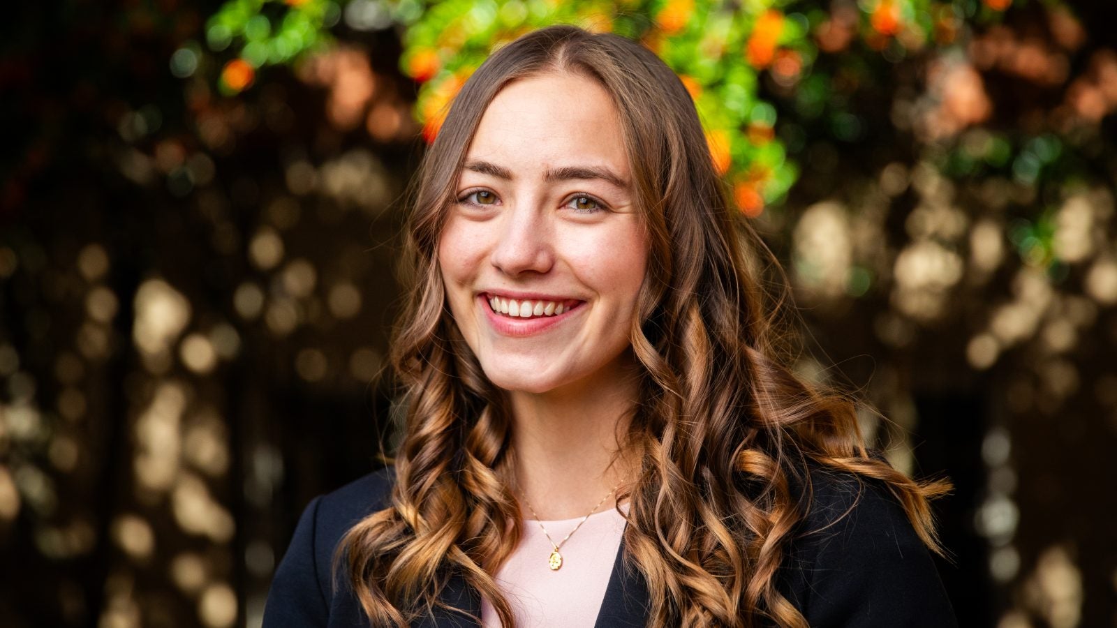 A white woman with brown curly hair smiles in front of fall leaves.