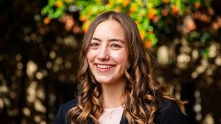 A white woman with brown curly hair smiles in front of fall leaves.